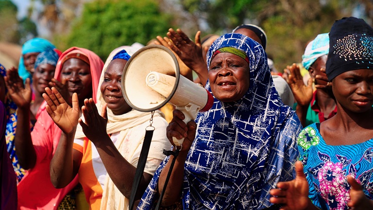 Groupe de femmes souriantes applaudissant en plein air, l'une au premier plan parlant dans un mégaphone, voile bleu à motifs