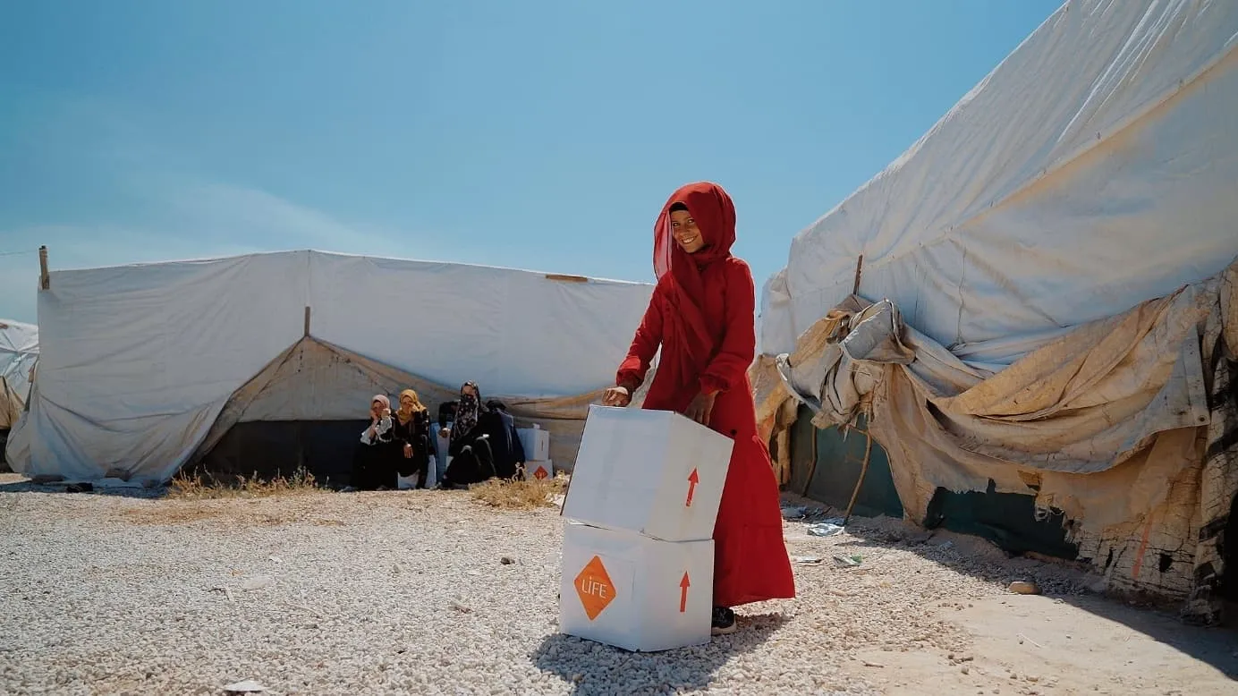 Une jeune fille en rouge sourit en portant un carton LIFE devant des tentes blanches usées d'un camp, des femmes assises derrière