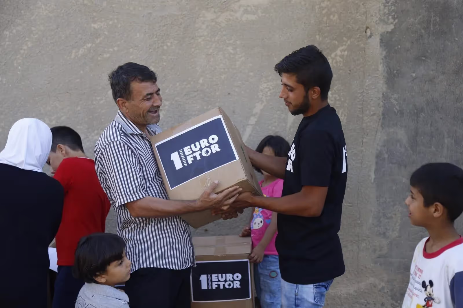 Un homme souriant reçoit un carton d'aide des mains d'un jeune bénévole, entouré d'enfants et de femmes, lors d'une distribution