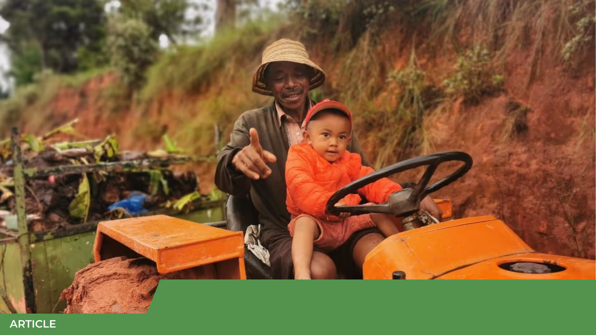 Un homme au chapeau de paille guide un jeune enfant en orange assis au volant d'un tracteur orange, sur un chemin de terre rouge