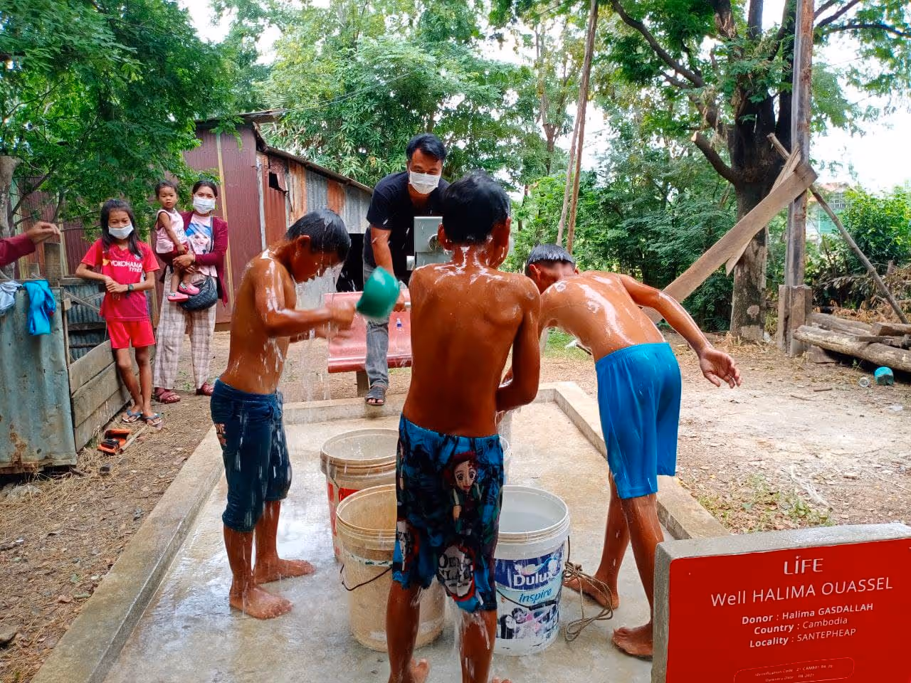 Trois garçons se lavant et se rinçant à un puits, avec des seaux ; panneau rouge LIFE indiquant Cambodia, Santepheap