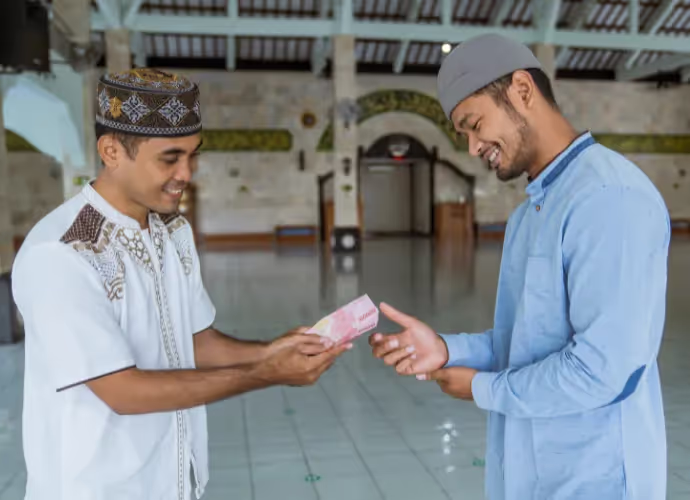 Deux hommes en tenue traditionnelle et calotte sourient dans une salle de mosquée, l'un tend des billets de banque à l'autre