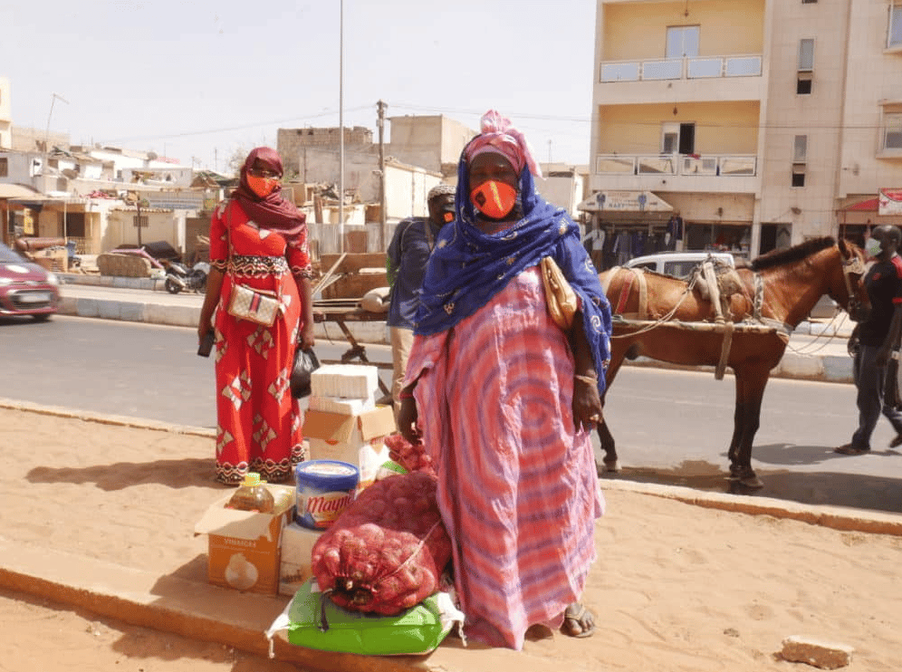 Deux femmes masquées dans une rue, avec des vivres : sac d'oignons, huile et cartons ; un cheval attelé derrière