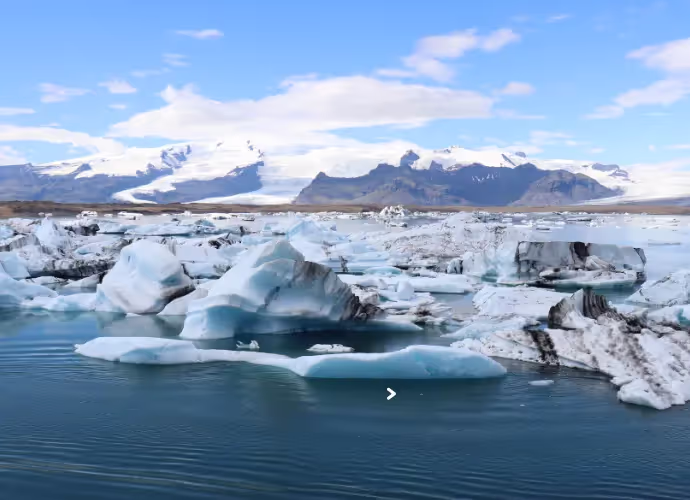 Icebergs blancs et bleutés flottant sur une eau calme, avec des montagnes enneigées et un ciel bleu en arrière-plan