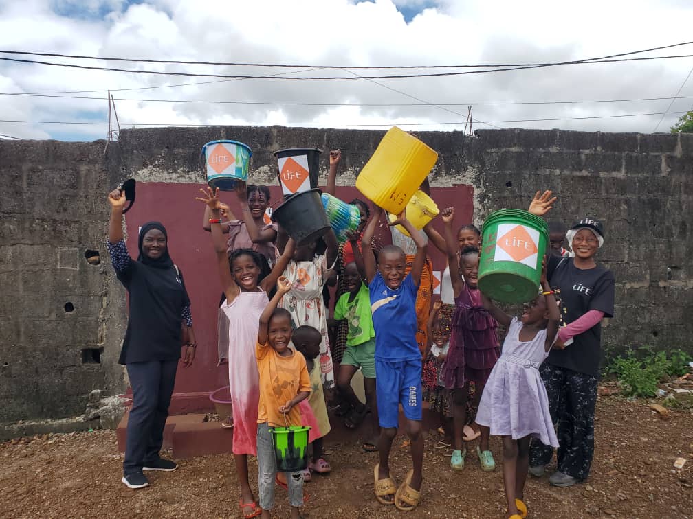 Enfants joyeux, bras levés, brandissant des seaux et bidons dont certains portent le logo LIFE, devant un mur en béton