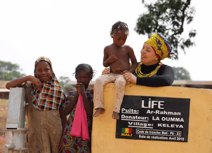 Enfants et une femme souriante posant près d'une pompe et d'une plaque LIFE du puits Ar-Rahman, village de Keleya, Mali