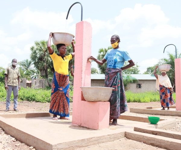 Des femmes remplissent des bassines à une fontaine rose, dans un village ; l'une porte une bassine sur la tête.