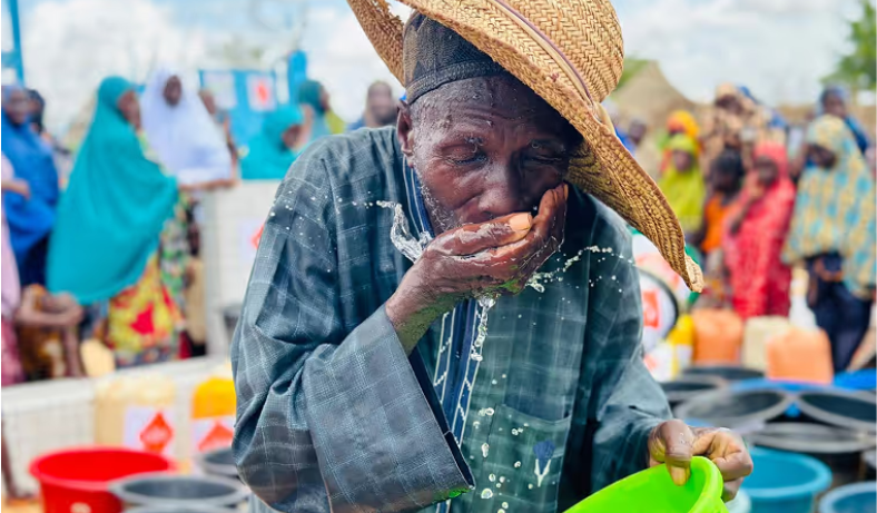 Un homme âgé coiffé d'un chapeau de paille boit de l'eau dans sa main, entouré de bidons et d'une foule de villageois