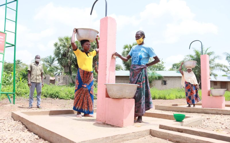 Deux femmes portant des bassines sur la tête ou à la main près de fontaines roses, sur une dalle en béton