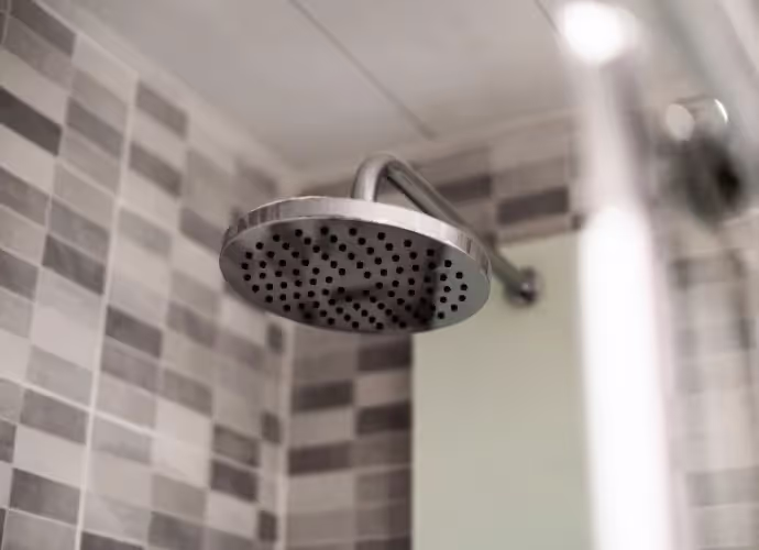 Pomme de douche ronde en métal fixée à un bras mural, dans une salle de bain aux murs carrelés de gris