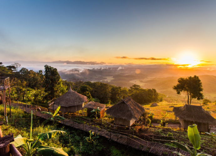 Lever de soleil sur des collines boisées couvertes de brume, avec des cabanes au toit de chaume au premier plan