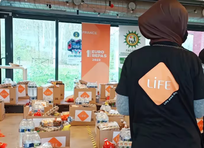 Bénévole de dos en t-shirt LIFE devant des tables de cartons de denrées et de bouteilles d'eau, lors d'une distribution alimentaire
