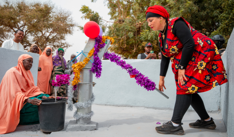 Une femme en tenue rouge à fleurs inaugure une pompe à eau décorée de guirlandes, une femme assise remplit un seau