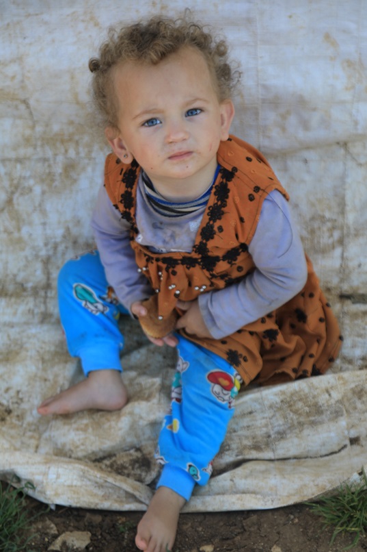 Un jeune enfant aux cheveux bouclés et pieds nus est assis sur une bâche sale et regarde l'objectif