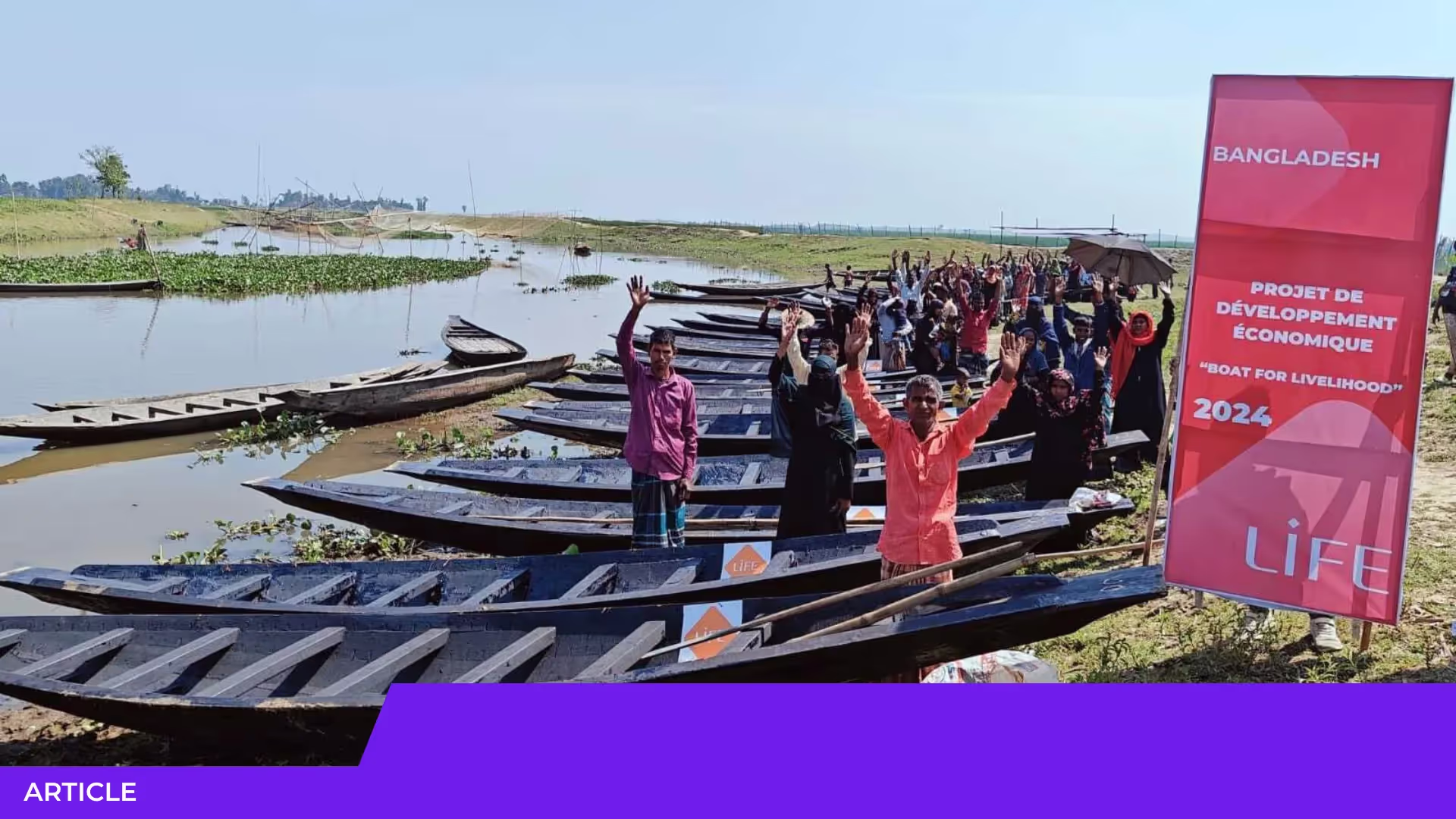 Groupe de villageois bras levés devant des barques alignées au bord de l'eau, près d'un panneau LIFE « Boat for livelihood » au Bangladesh