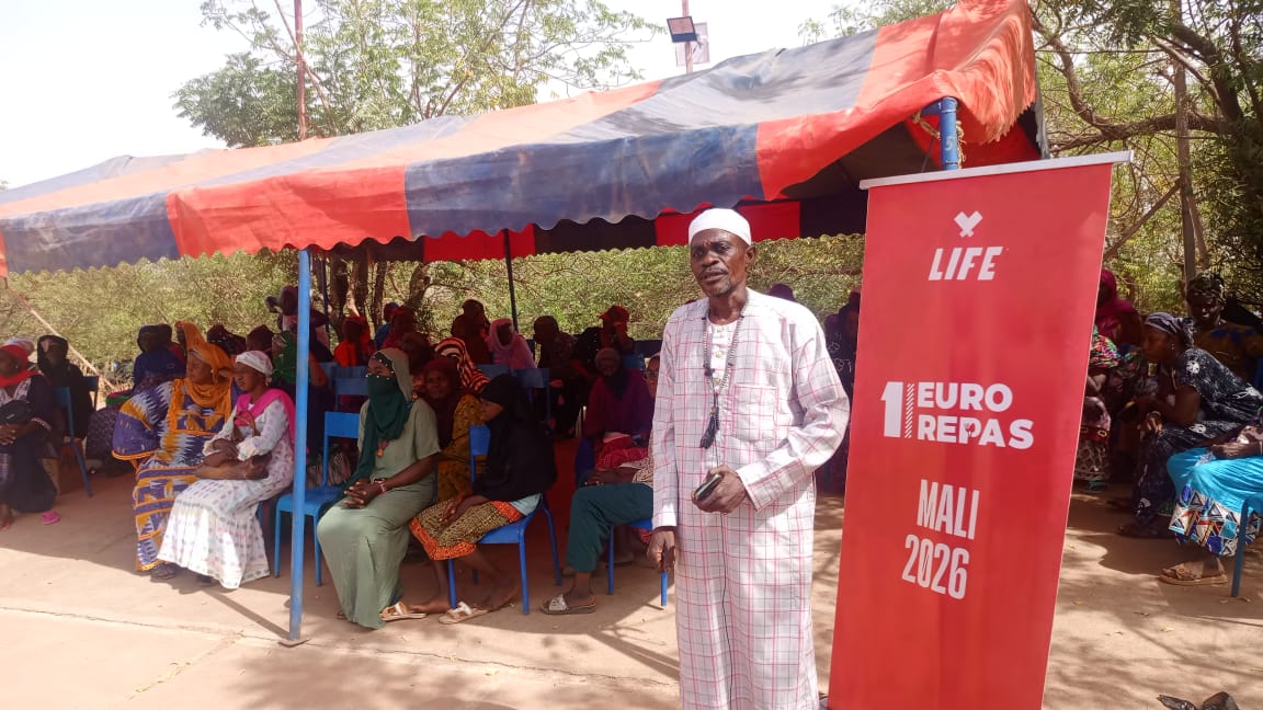 Un homme debout en tenue claire devant une bannière LIFE « 1 Euro Repas Mali 2026 », face à des femmes assises sous une bâche