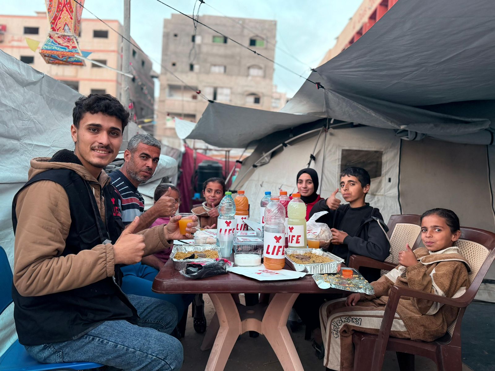 Repas partagé à une table sous des tentes : un bénévole et des enfants sourient, pouce levé, devant des bouteilles et barquettes LIFE.