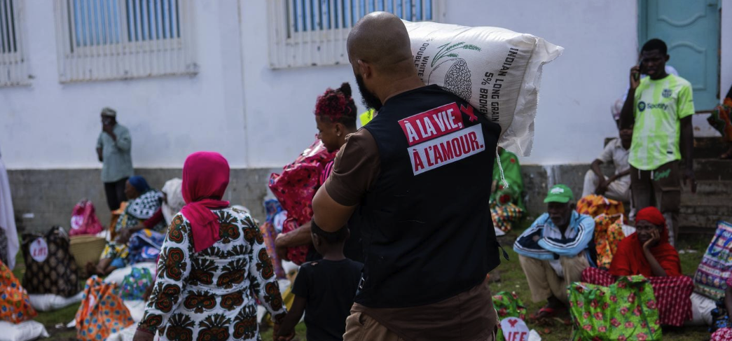 Un bénévole en gilet noir « À la vie, à l'amour » porte un sac de riz sur l'épaule devant des familles assises lors d'une distribution
