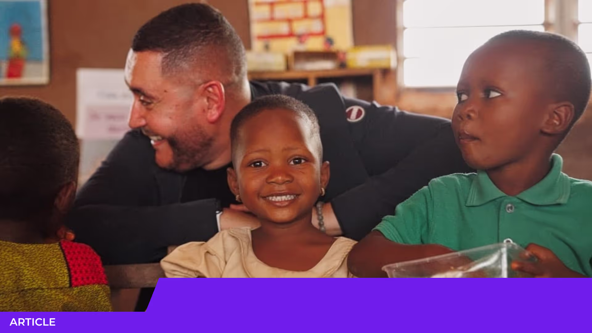 Homme souriant penché derrière une petite fille souriante, dans une salle de classe, avec deux autres enfants dont un garçon en polo vert.