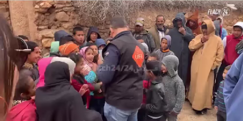 Un homme portant un gilet LIFE distribue quelque chose à des enfants entourés d'habitants d'un village de pierre