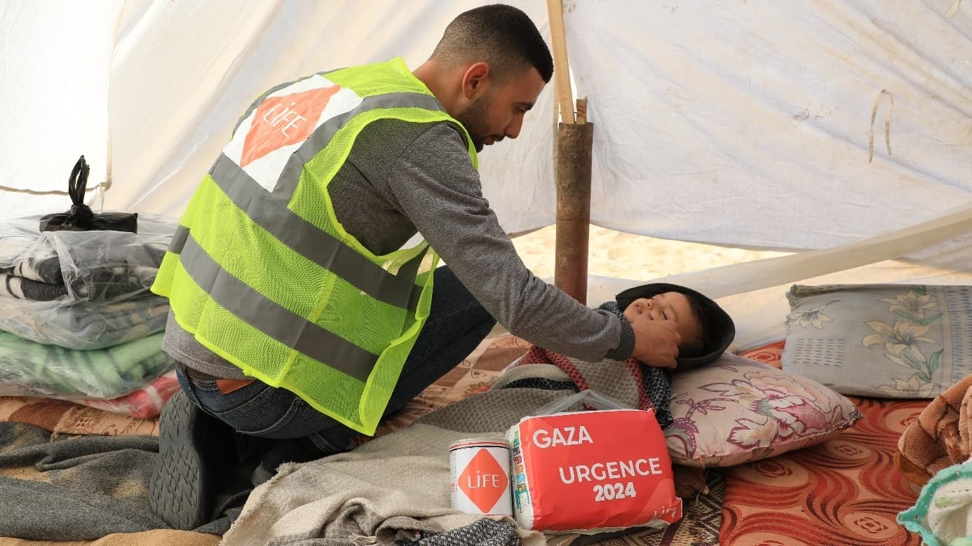 Un bénévole en gilet jaune LIFE se penche vers un enfant allongé sous une tente, avec un colis « Gaza Urgence 2024 »