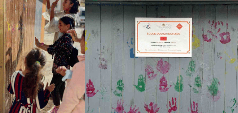 Des enfants laissent des empreintes de mains colorées sur une palissade en bois, avec une plaque indiquant « École Douar Inghade » et un drapeau.