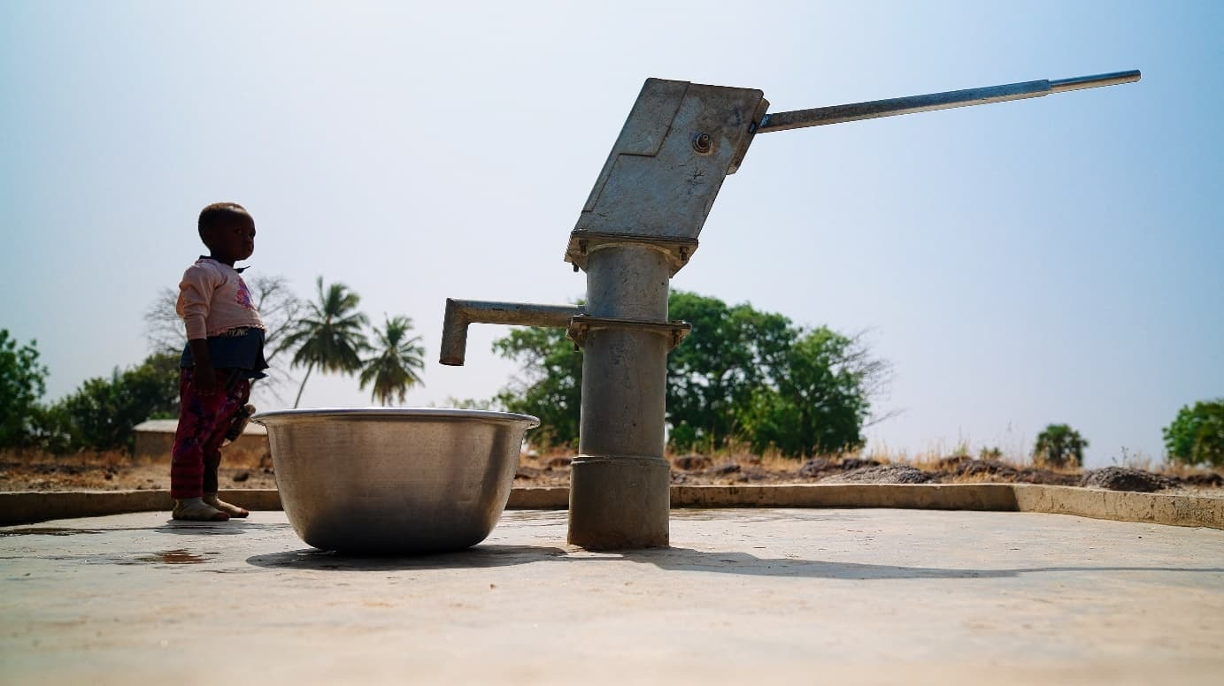 Un jeune enfant debout près d'une pompe à eau manuelle et d'une grande bassine en métal, sous un ciel clair