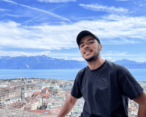 Jeune homme souriant, casquette et t-shirt noirs, posant devant une ville aux toits rouges, un lac et des montagnes