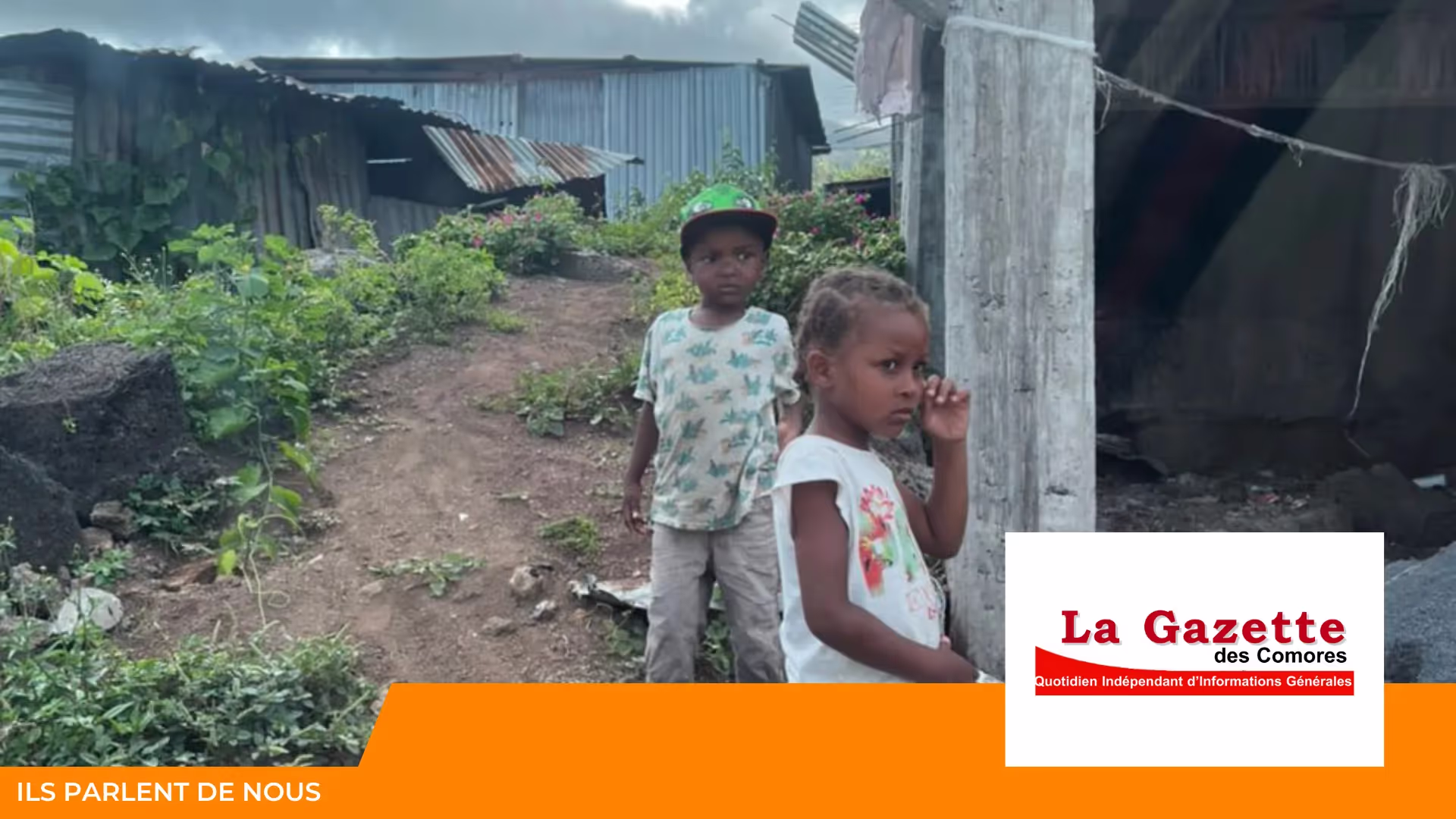 Deux jeunes enfants debout sur un chemin de terre devant des maisons en tôle, avec le logo de La Gazette des Comores