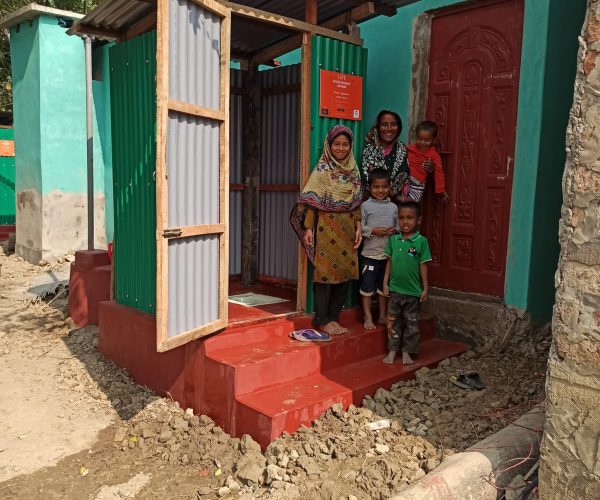 Une femme et trois enfants souriants posent sur les marches rouges d'une latrine en tôle verte construite par LIFE