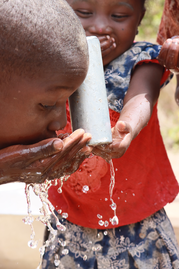 Gros plan sur un enfant qui boit de l'eau dans ses mains au bec d'une pompe, un autre enfant en rouge derrière lui