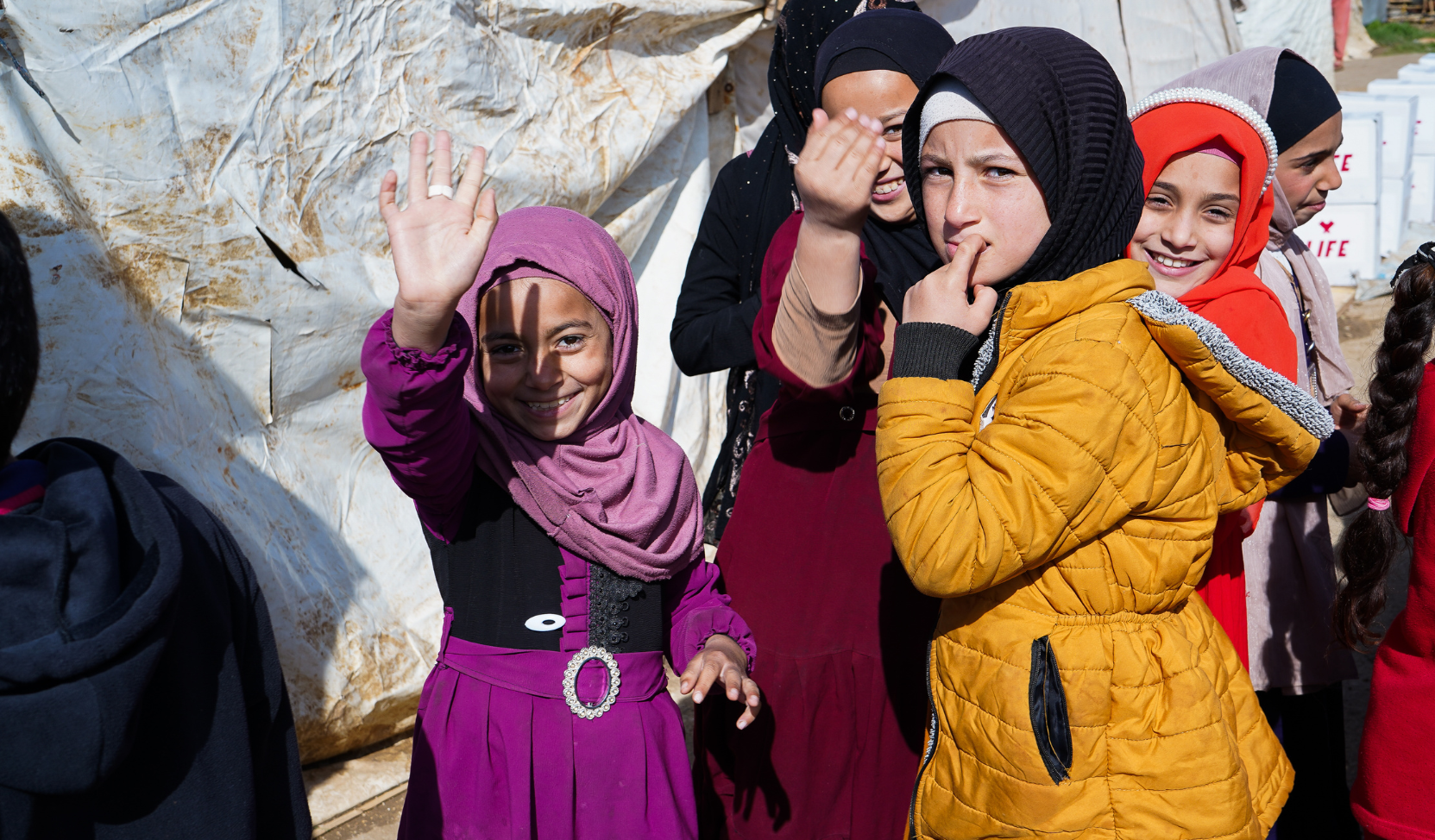 Fillettes souriantes en hijab et manteaux colorés, l'une faisant un signe de la main, devant une tente en bâche usée