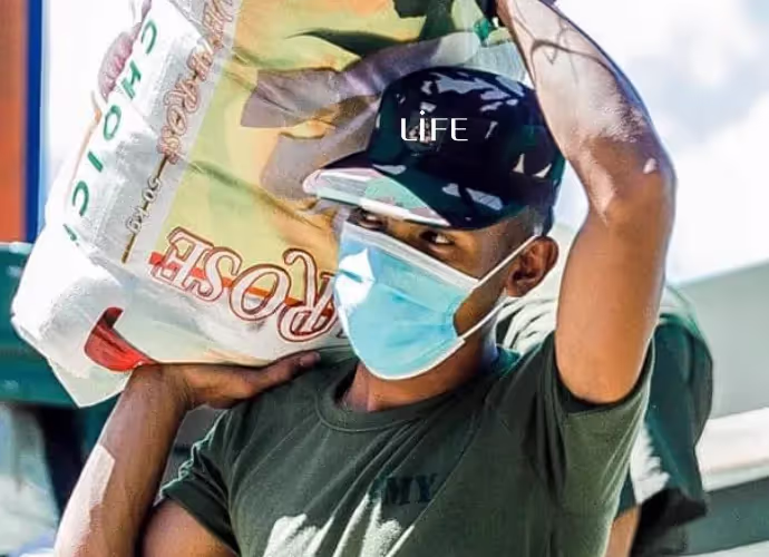 Homme portant une casquette LIFE et un masque chirurgical, un sac de riz sur l'épaule, bras levé