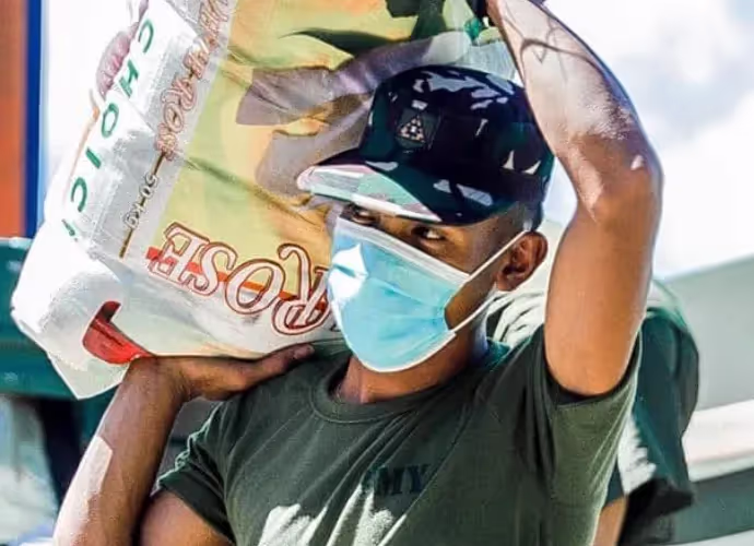 Homme portant un sac de riz sur l'épaule, casquette et masque chirurgical bleu, t-shirt vert militaire