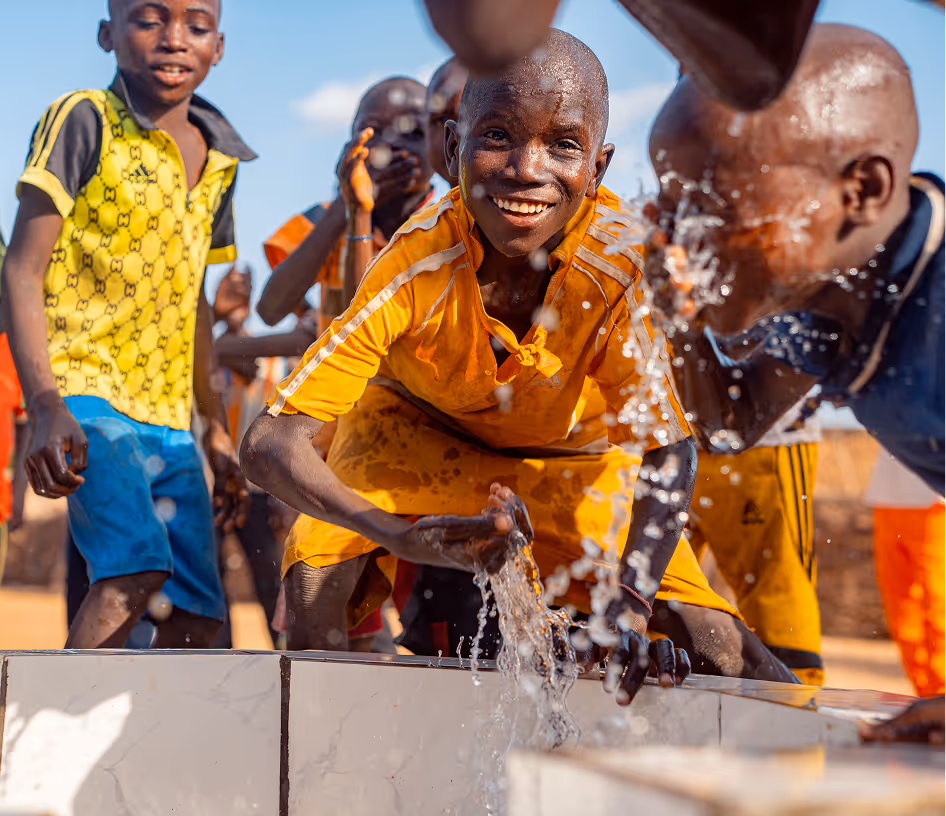 Un garçon en maillot jaune sourit en jouant avec l'eau qui coule d'un point d'eau, d'autres enfants rient derrière lui.