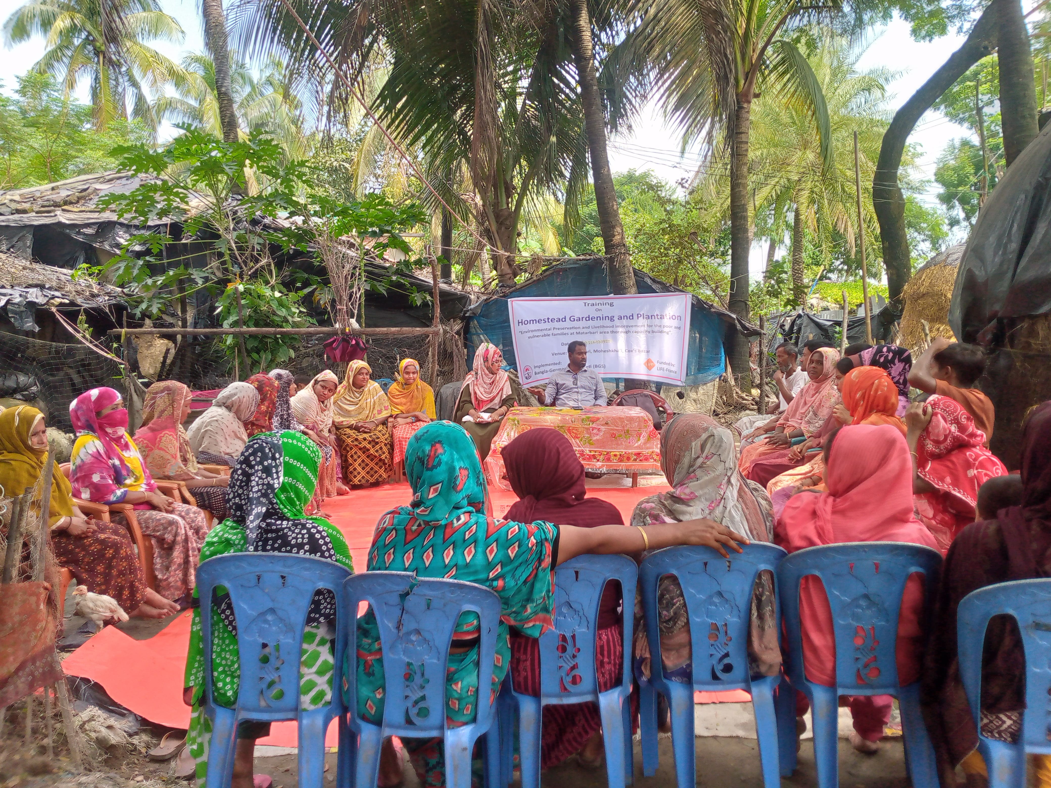 Formation en plein air : des femmes assises sur des chaises en plastique écoutent un animateur devant une banderole « Homestead Gardening and Plantation »
