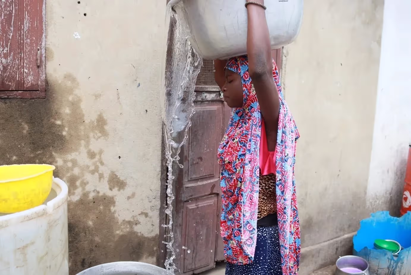 Femme portant sur la tête une grande bassine d'où l'eau se déverse, devant un mur et une porte en bois, des récipients à côté