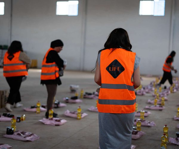 Des bénévoles LIFE en gilet orange préparent des colis alimentaires avec bouteilles d'huile dans un grand entrepôt