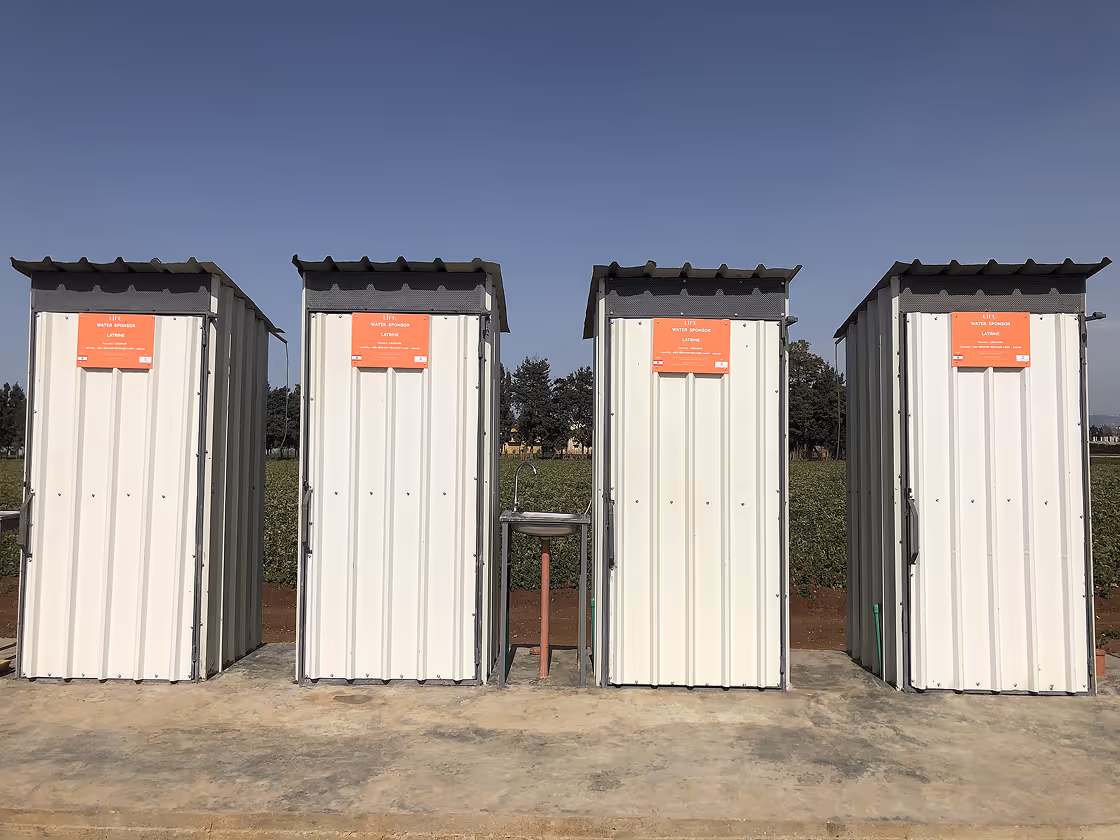 Quatre latrines blanches en tôle avec une affiche orange LIFE, un lavabo entre elles, devant un champ cultivé.