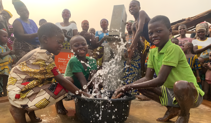 Des enfants souriants s'éclaboussent d'eau autour d'une pompe et d'une bassine, sous le regard d'une foule de villageois