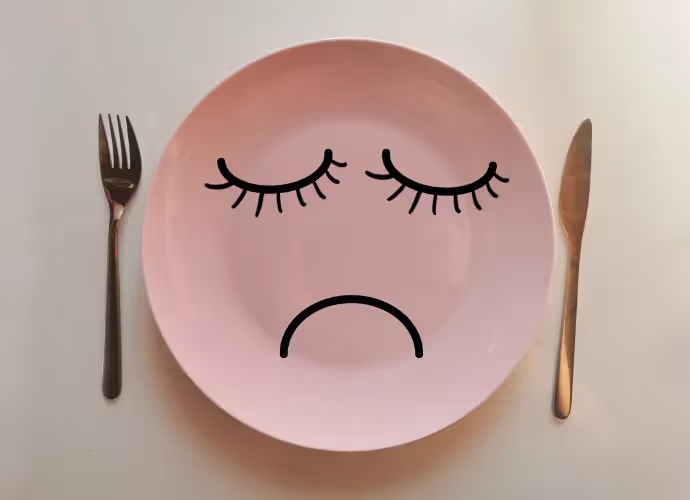Assiette rose vide avec une fourchette et un couteau, sur laquelle sont dessinés des yeux fermés et une bouche triste