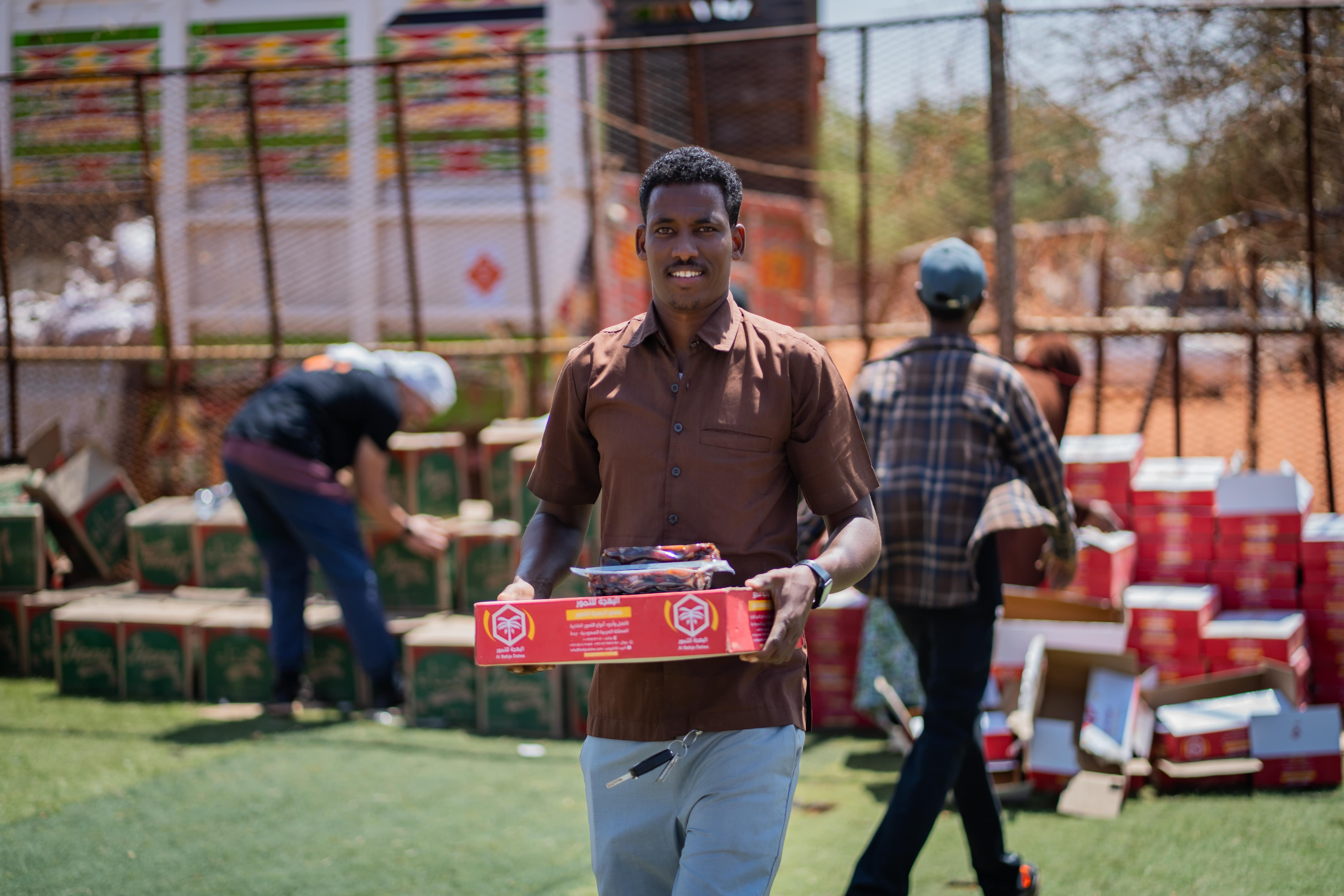 Un homme souriant porte un carton rouge de denrées devant des piles de cartons rouges et verts sur un terrain d'herbe