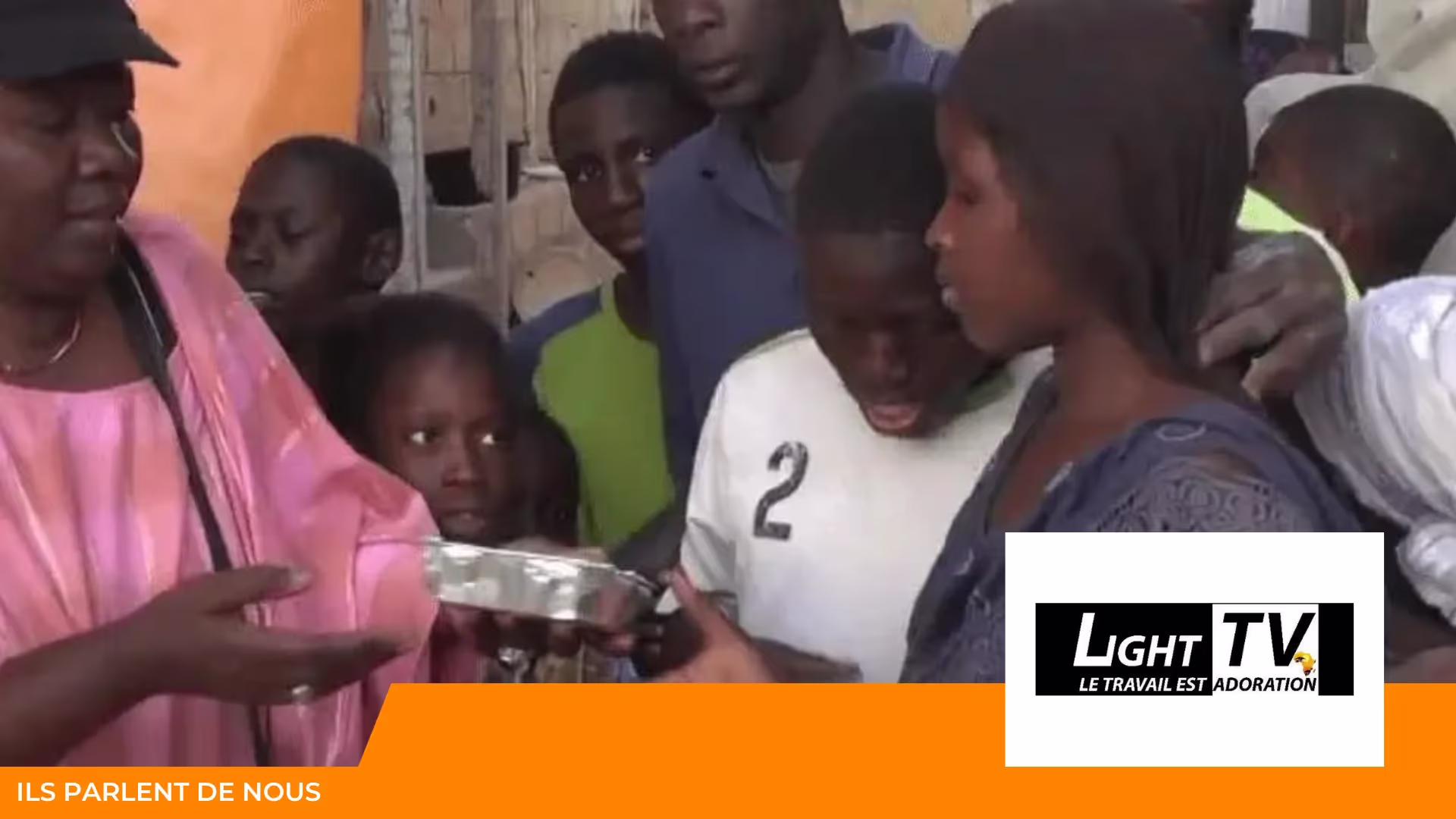 Une femme en tenue rose tend une barquette en aluminium à des enfants et des jeunes ; logo Light TV, « Ils parlent de nous »