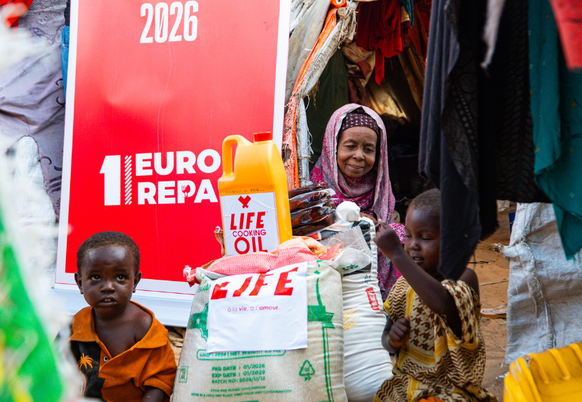 Deux enfants et une femme âgée devant une bannière « 1 Euro Repas », avec un sac de riz LIFE et un bidon d'huile LIFE Cooking Oil
