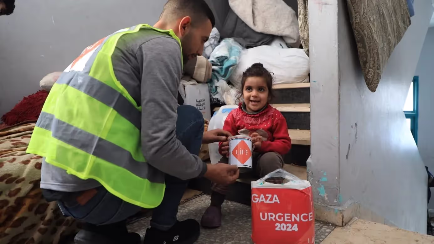 Un bénévole en gilet jaune fluo tend une boîte LIFE à une petite fille en rouge assise dans un escalier, colis « Gaza Urgence 2024 »