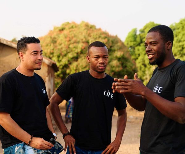 Trois hommes en t-shirt noir LIFE discutent en plein air dans un village, l'un d'eux parle en gesticulant