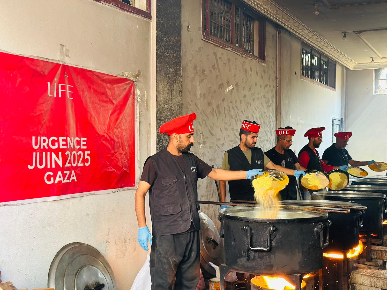 Des bénévoles LIFE coiffés de bérets rouges versent de la nourriture cuite dans des plats, devant une affiche « Urgence juin 2025 Gaza »