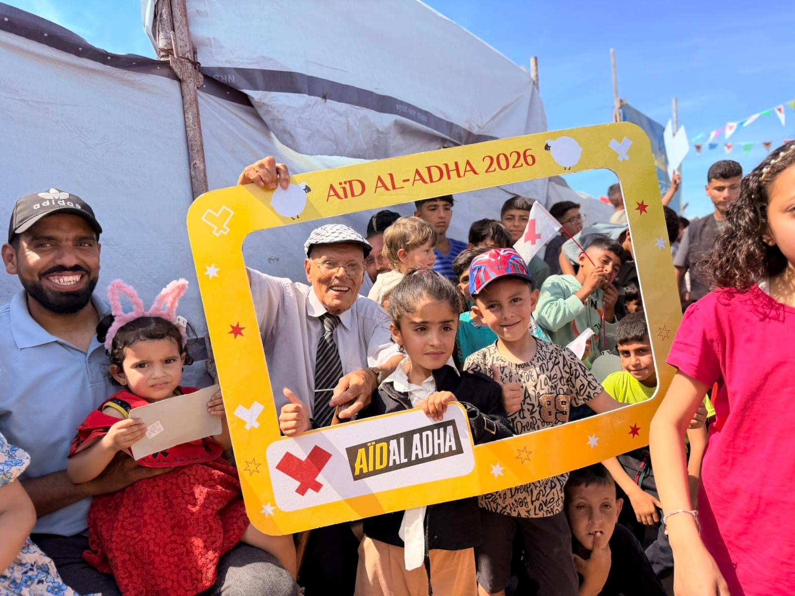 Enfants et adultes souriants derrière un cadre photo « Aïd al-Adha 2026 » devant une tente, sous un ciel bleu