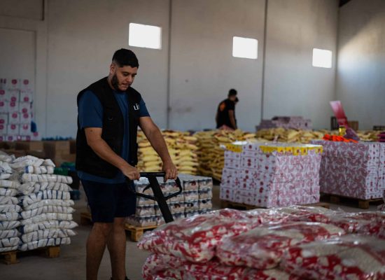 Un homme en gilet noir LIFE tire un transpalette dans un entrepôt rempli de palettes de denrées alimentaires