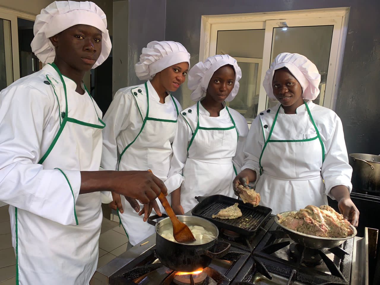 Quatre apprentis en tenue de cuisinier blanche à liseré vert cuisent de la viande et préparent un plat sur une cuisinière.